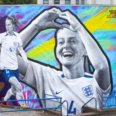 Large mural on brick wall of female footballer in England shirt making a love heart with her hands on a colourful background