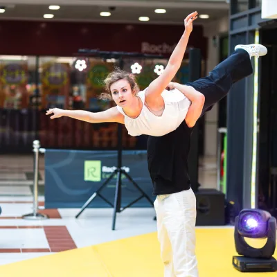 Woman in white top, black trousers and white shoes being held up by person in black top and white trousers with seated audience on yellow floor in shopping centre